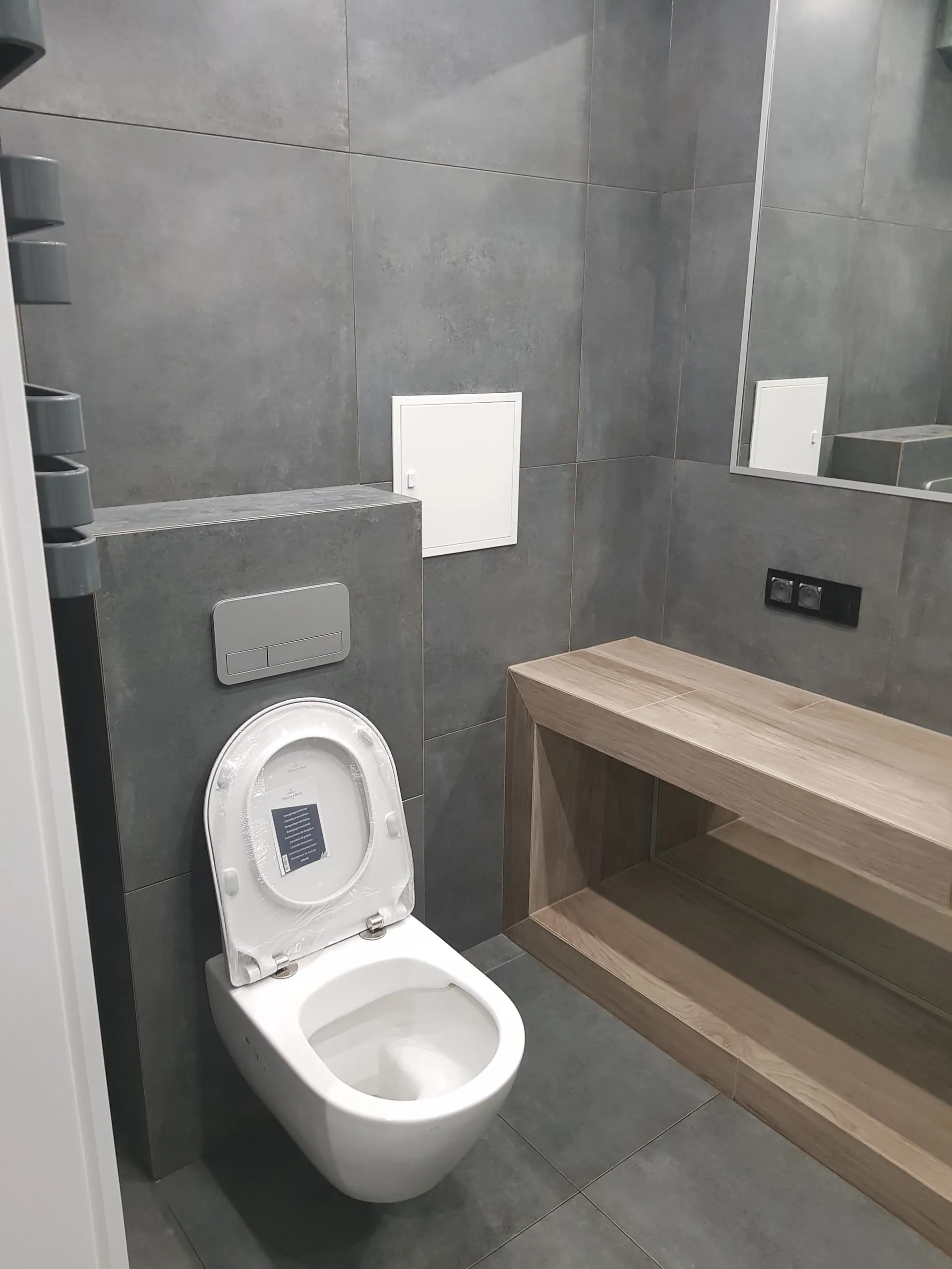Strefa WC z podwieszaną miską, szarą zabudowa stelaża i drewnianą ławką z półką.